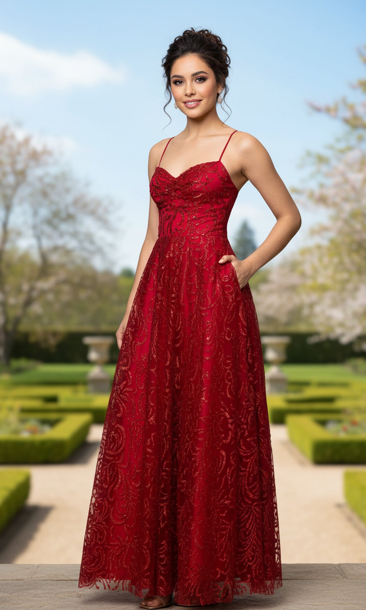 A woman stands outdoors in a manicured garden wearing the Blondie Nites 6101BN long prom dress—a sleeveless, floor-length red gown with lace detailing, thin straps, and elegant charm.